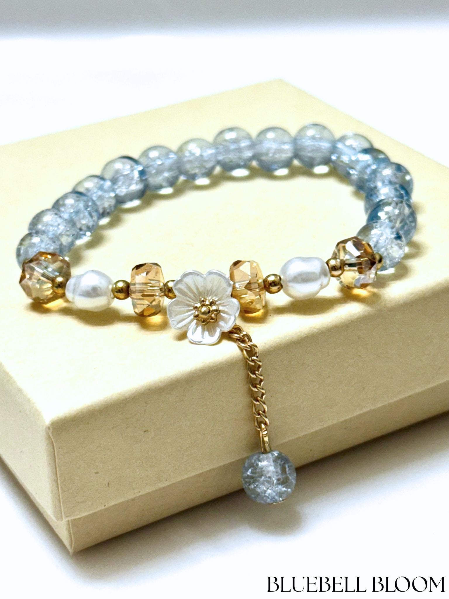 Handmade beaded bracelet with blue crackle beads, white flower charm, and gold crystal accents on a beige box
