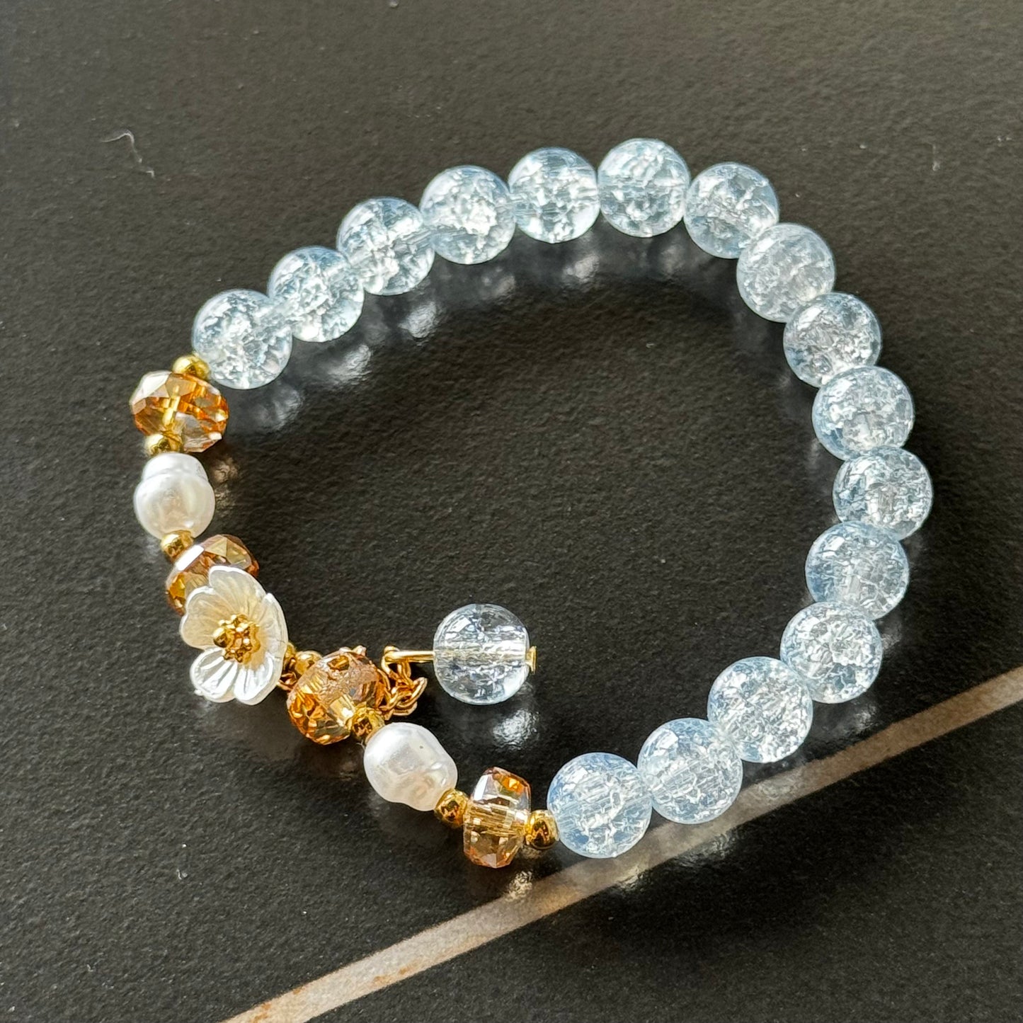 Handmade beaded bracelet with blue crackle beads, white flower charm, and gold crystal accents on a beige box