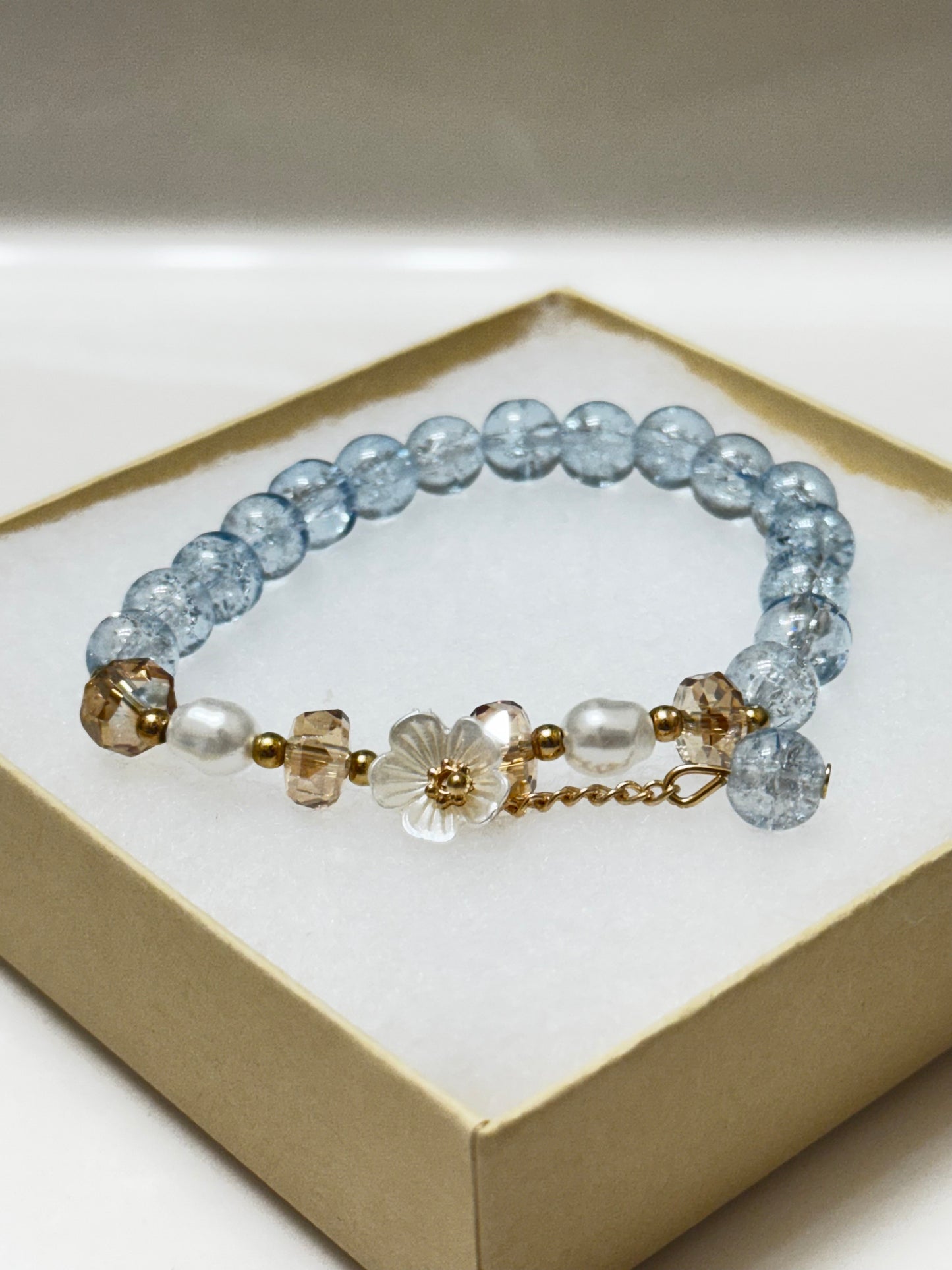 Handmade beaded bracelet with blue crackle beads, white flower charm, and gold crystal accents on a beige box