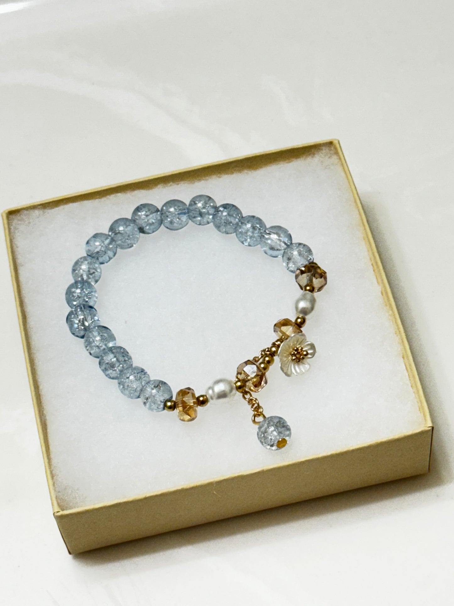 Handmade beaded bracelet with blue crackle beads, white flower charm, and gold crystal accents on a beige box