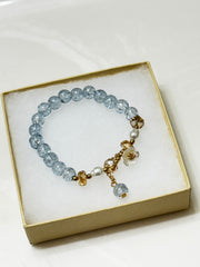 Handmade beaded bracelet with blue crackle beads, white flower charm, and gold crystal accents on a beige box