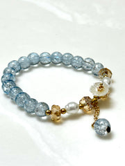 Handmade beaded bracelet with blue crackle beads, white flower charm, and gold crystal accents on a beige box