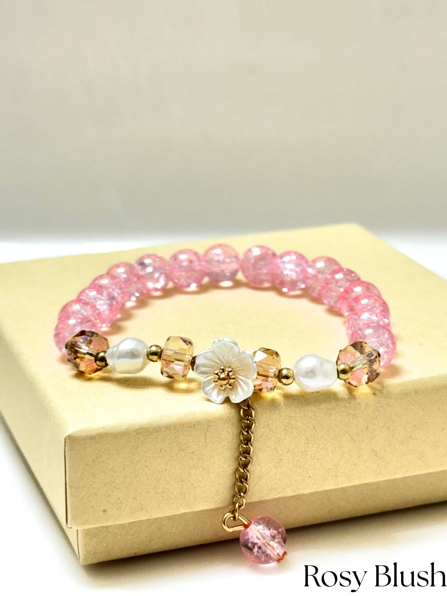 Handmade pink crackle beaded bracelet with white flower charm, gold crystals, and dangling bead, displayed in a gift box